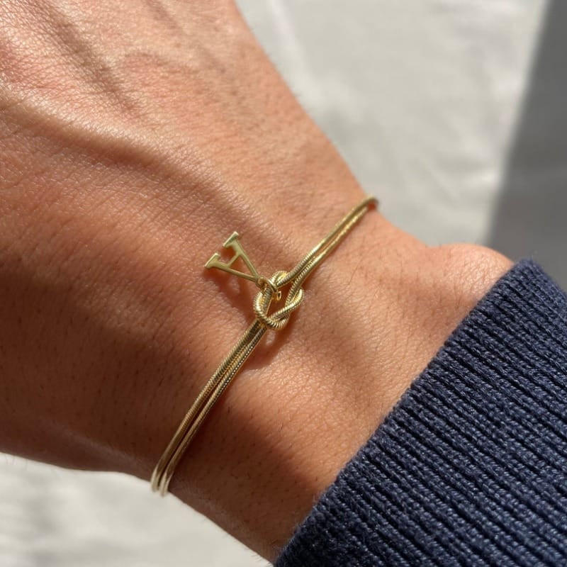 A close-up of a wrist wearing the Bracelet with love knot and letters, featuring an abstract intertwined design. The bracelet is paired with a dark, ribbed long-sleeve shirt against a softly blurred background.