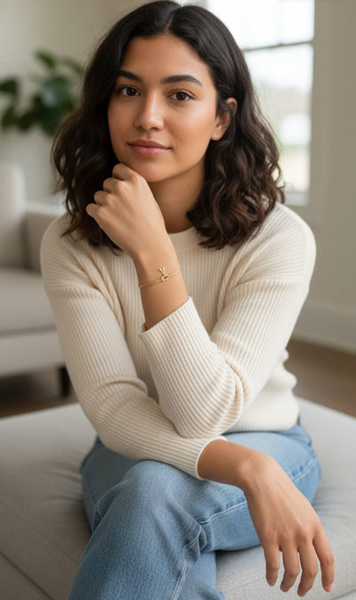 A young woman with wavy dark hair sits indoors on a light-colored couch, wearing a cream sweater, light blue jeans, and the Bracelet with love knot and letters. She rests her chin on her hand and gazes calmly at the camera.